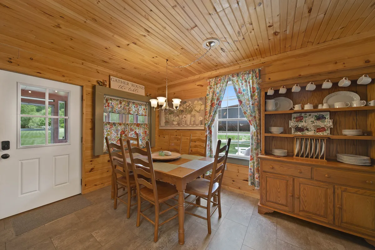 A bright, cozy dining nook featuring a wooden dining table, floral curtains, a hutch with dishware, and a large window letting in natural light. The ambiance is welcoming and ideal for coffee moments or evening meals with loved ones.