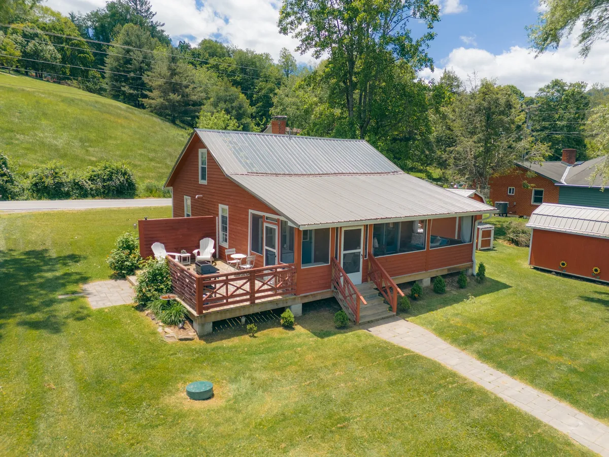 This image features the red exterior of Broadstone Cottage, a welcoming deck with comfortable chairs, a manicured lawn, and lush surrounding greenery. The deck provides an inviting spot for relaxation or social gatherings, while the greenery and open space hint at the tranquil, walkable neighborhood setting.