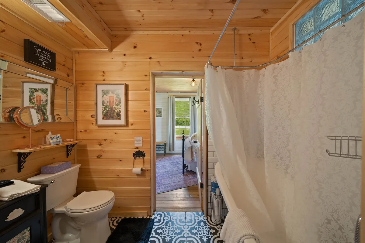 A charming bathroom with wooden walls, a decorative shower curtain, a clean sink area, framed artwork, and patterned floor tiles, evoking a cozy, cottage-like feel while ensuring essential comfort.
