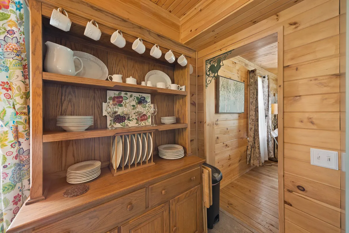 A cozy kitchen area featuring a wooden hutch filled with kitchenware, colorful curtains adding cheer, and a clear view through a doorway into an adjacent room. The space feels warm and inviting, perfect for preparing meals and gathering together.