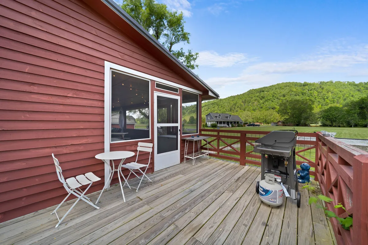 A casual, relaxed exterior view of the home's red wooden facade, spacious deck area, outdoor seating, and a grill. The deck offers a perfect spot for outdoor dining, relaxing with friends, or taking in the natural beauty of the mountains. Lush greenery surrounds the home, reinforcing the year-round appeal of the setting.