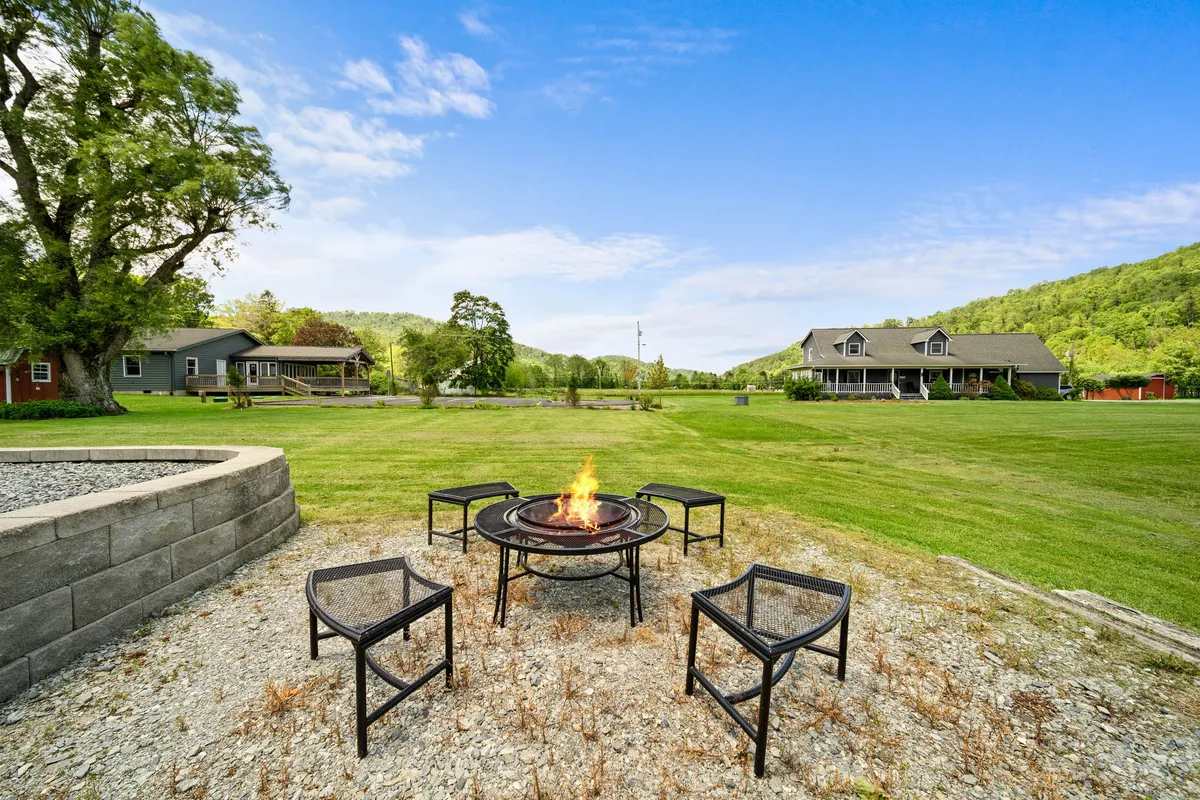 A cozy fire pit area surrounded by comfortable chairs sits on a green lawn with a sweeping mountain backdrop. The scene is set for evening gatherings, with the fire pit ready for marshmallows and stories under the open sky.