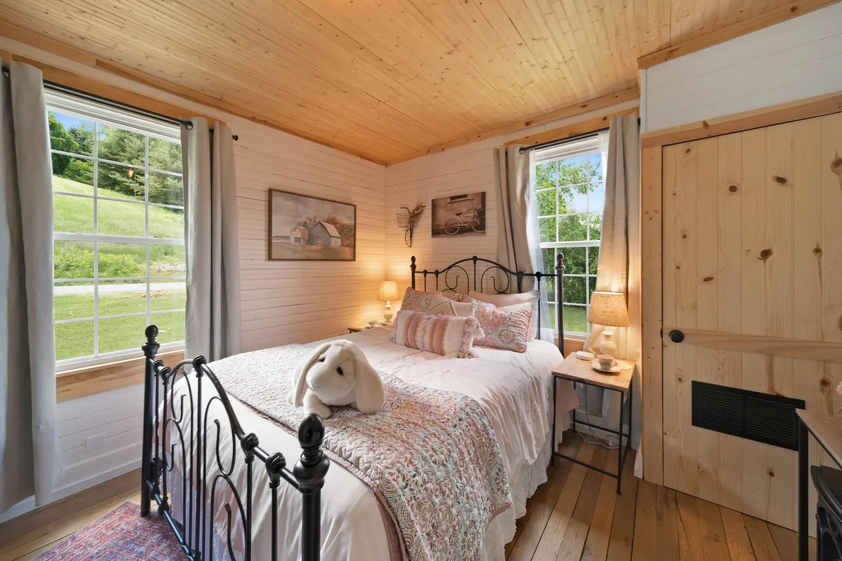 A tranquil bedroom with a window that lets in natural light, a bed layered with decorative pillows, a plush white bunny for a playful touch, uplifting artwork, and a nightstand with a lamp—creating a restful, storybook-like atmosphere.