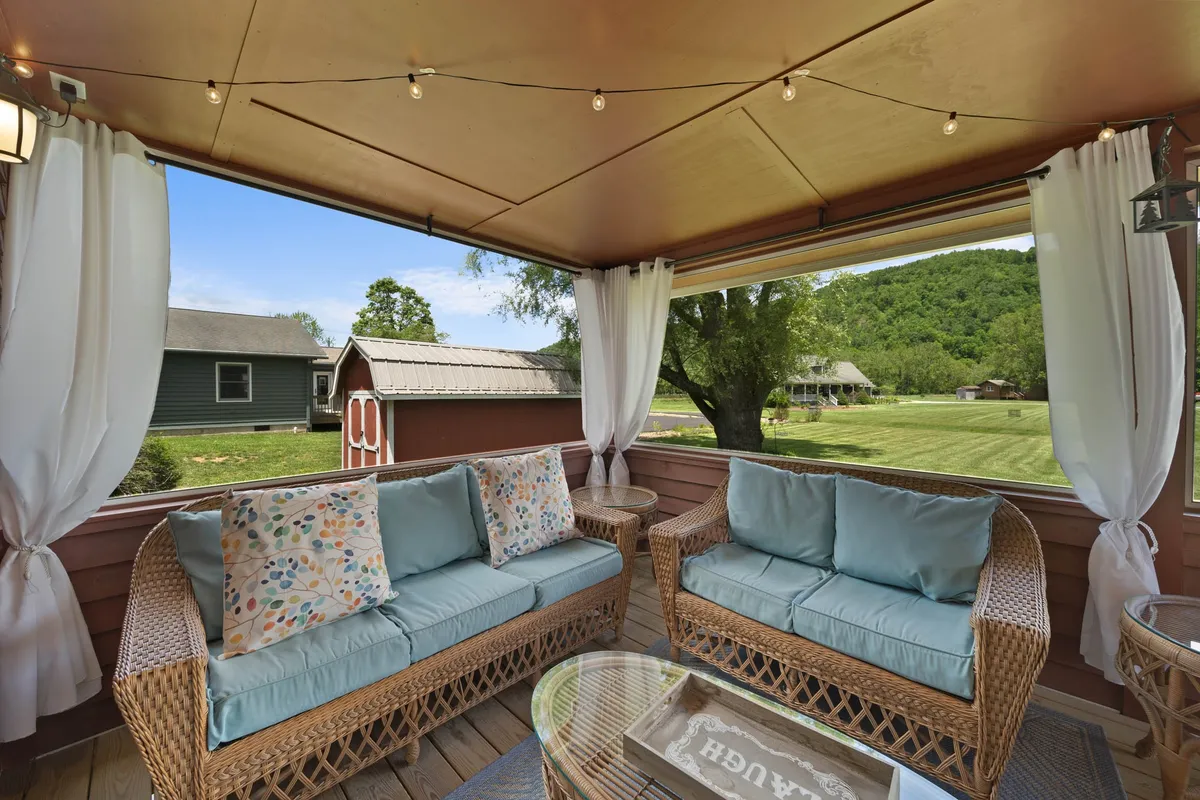 A covered porch with woven furniture and decorative cushions is softly illuminated by string lights, offering a peaceful view of the landscape. The cozy setup invites relaxation, whether for morning coffee or enjoying the sunset.