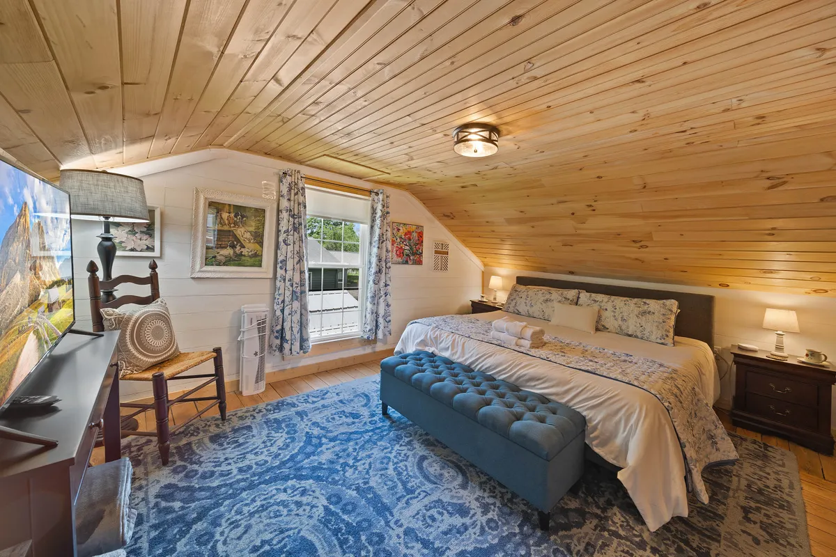 A serene upstairs bedroom with a spacious bed covered in decorative bedding, a tufted bench at the foot, a television, wooden chair, and light streaming in through large windows with soft curtains—providing a sense of comfort beneath potentially sloped ceilings.